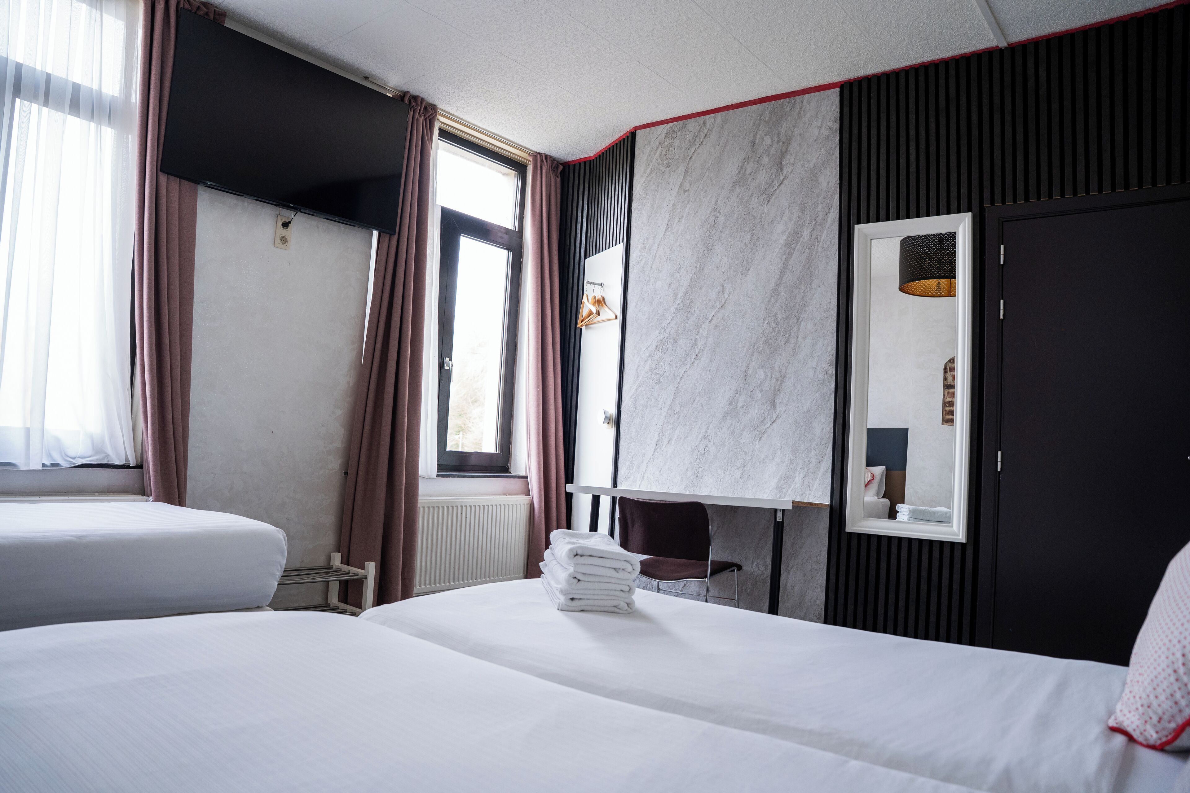 Hotelkamer met twee bedden, een raam met gordijnen, een radiator, een spiegel en een stapel handdoeken.