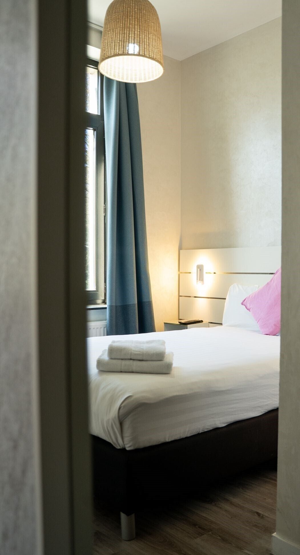 Una habitación de hotel con una cama, una mesita de noche, una lámpara y una ventana con cortinas.
