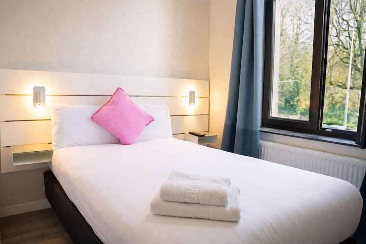 A neatly made bed with white linens and a pink pillow, bedside lamps, and a window with a view of trees.