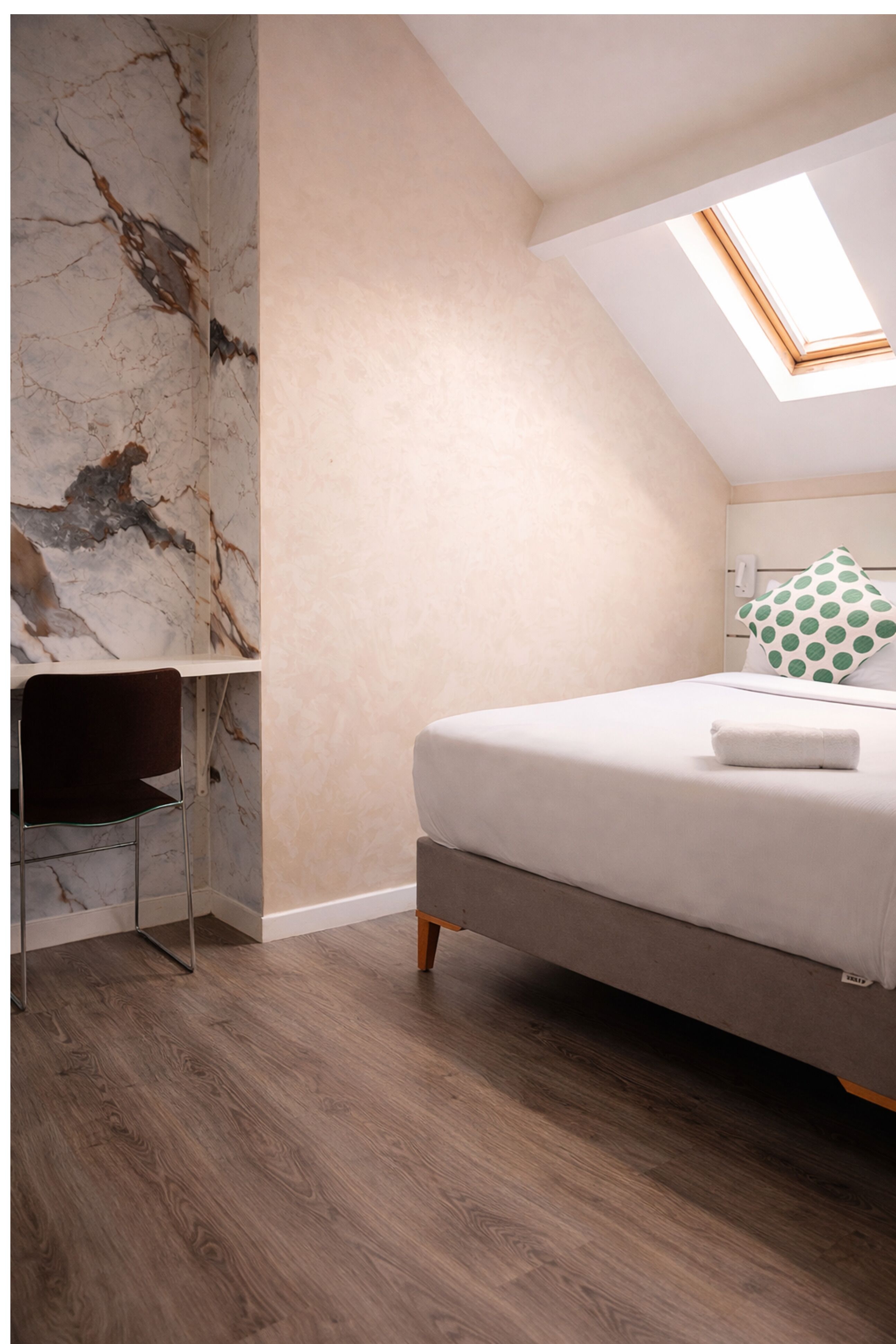 Une chambre à coucher avec un lit, un bureau et une chaise. Il y a une lucarne au-dessus du lit.