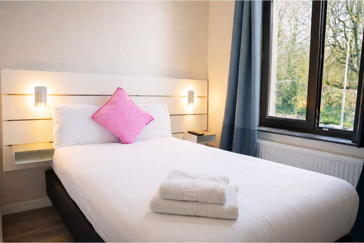 A neatly made bed with white linens and a folded white towel, a pink pillow, and two bedside lamps on a nightstand.
