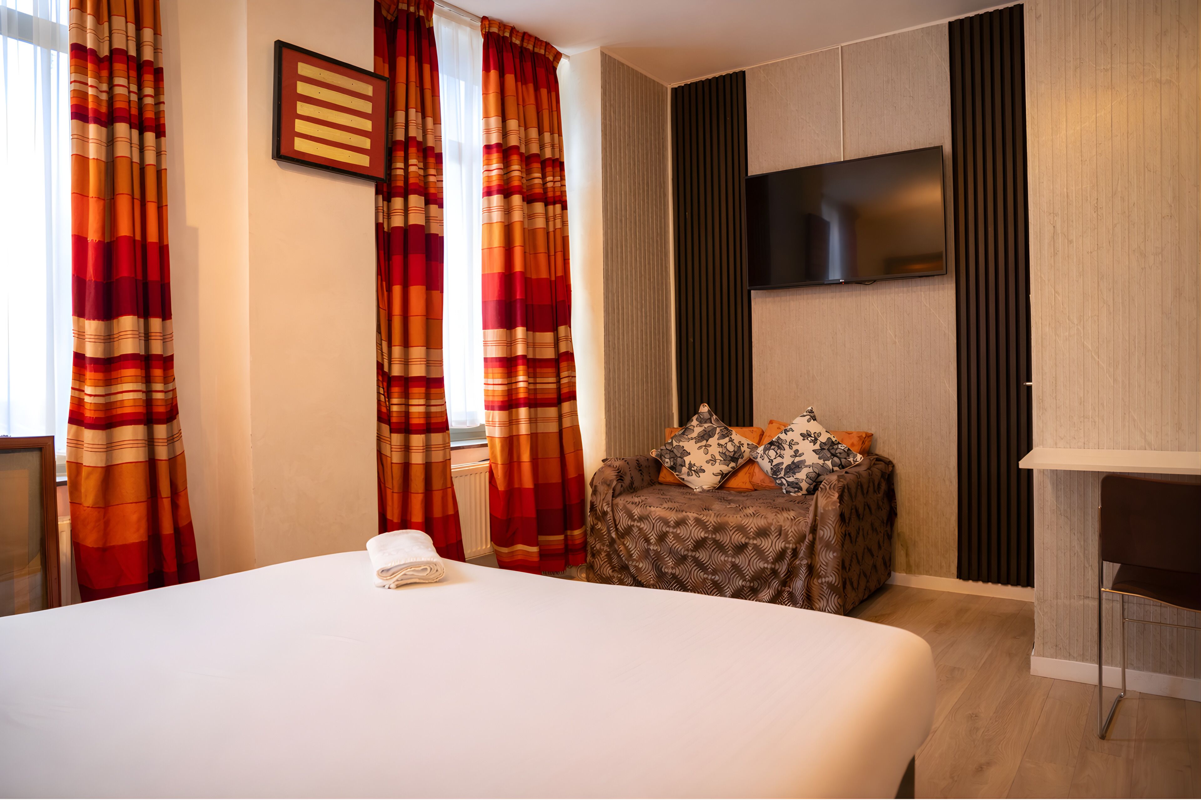 A hotel room with a large bed, a small table with decorative items, a flat-screen TV mounted on the wall, and striped curtains.