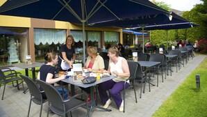 An outdoor dining area with tables and chairs under a large umbrella, surrounded by a building with windows and a garden. - Hotel Garni Geisler (Cologne)