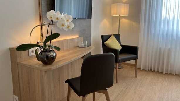 A modern hotel room with a wooden desk, a black chair, a vase with white orchids, a flat-screen TV, and a standing lamp. - Hotel Garni Geisler (Cologne)