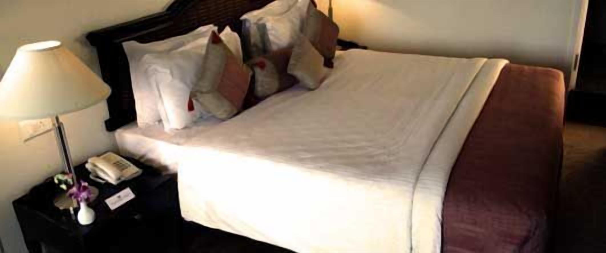 Una cama bien hecha con ropa de cama blanca y una manta marrón, una lámpara de noche y una mesita con un teléfono y un florero.