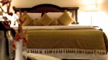A neatly made bed with a green bedspread and multiple pillows, a bedside table with a lamp, and a vase with flowers.