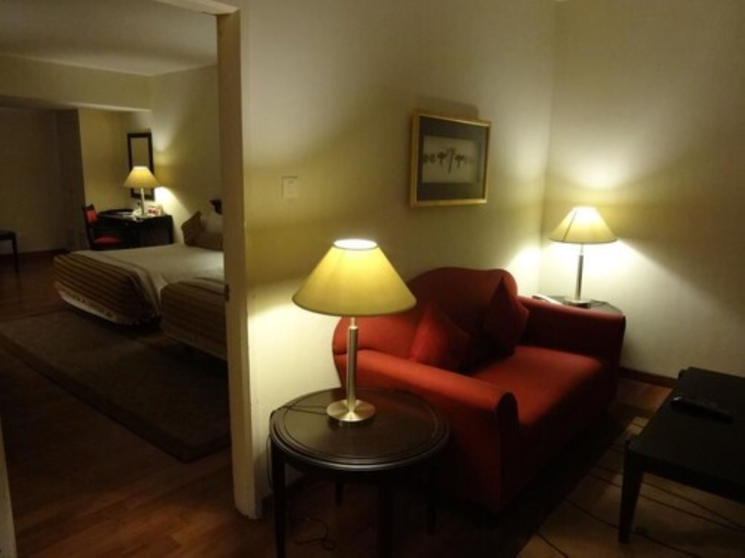 Habitación de hotel con cama, sofá rojo, una mesita con lámpara y un cuadro en la pared.
