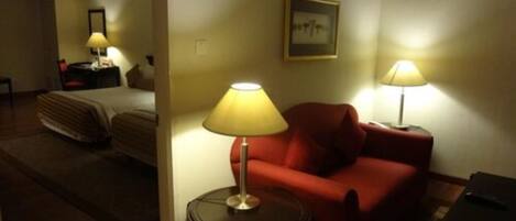 Habitación de hotel con cama, sofá rojo, una mesita con lámpara y un cuadro en la pared.