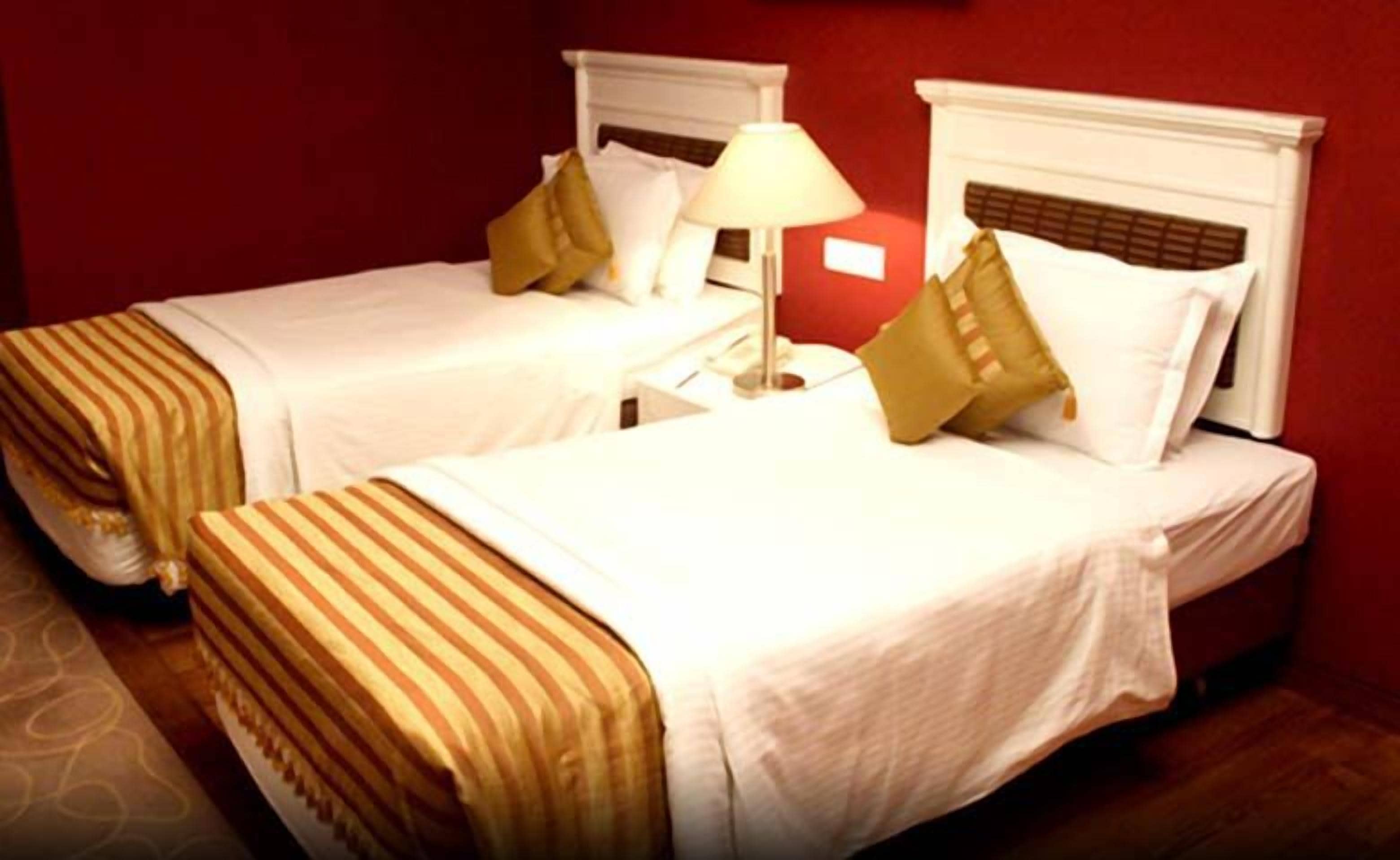 Two single beds with white headboards and beige bedding, a nightstand with a lamp, and a red wall.