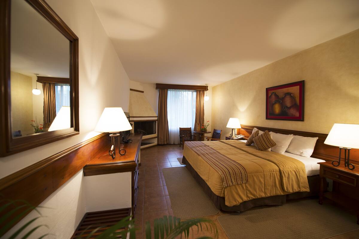 A hotel room with a large bed, two bedside tables with lamps, a desk with a chair, a large mirror, and a painting on the wall.
