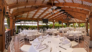 Ein Essbereich im Freien mit runden Tischen, gedeckt für eine festliche Veranstaltung, umgeben von weißen Stühlen und überdacht von einer Holzpergola.