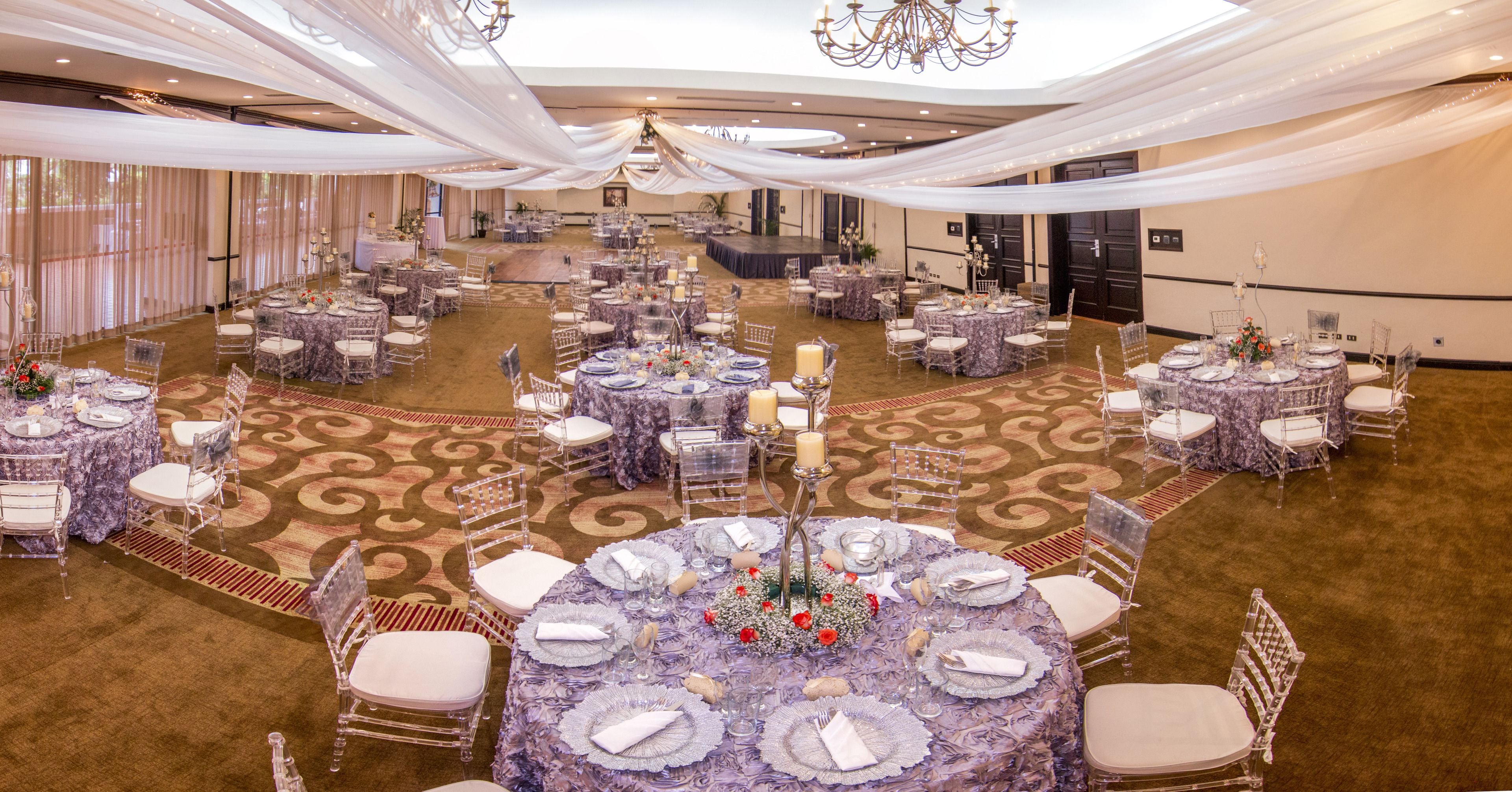 A banquet hall with round tables set for a formal event, featuring clear chairs and white tablecloths.