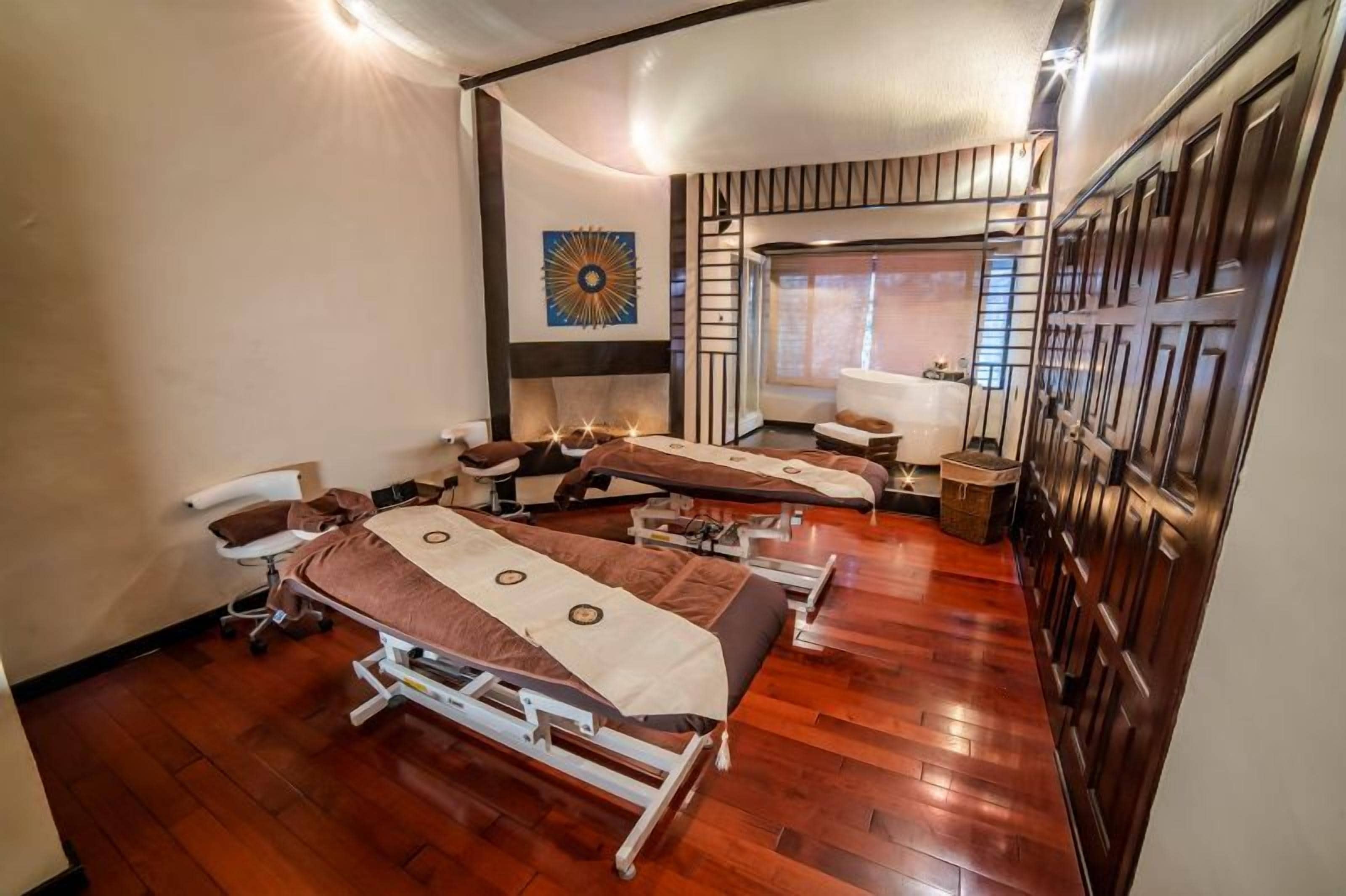 A room with three massage tables arranged in a row, a wooden floor, and a large window with blinds.