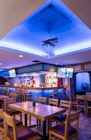 Una zona de bar con mesas y sillas de madera, una barra con un barman y dos televisores montados en la pared.