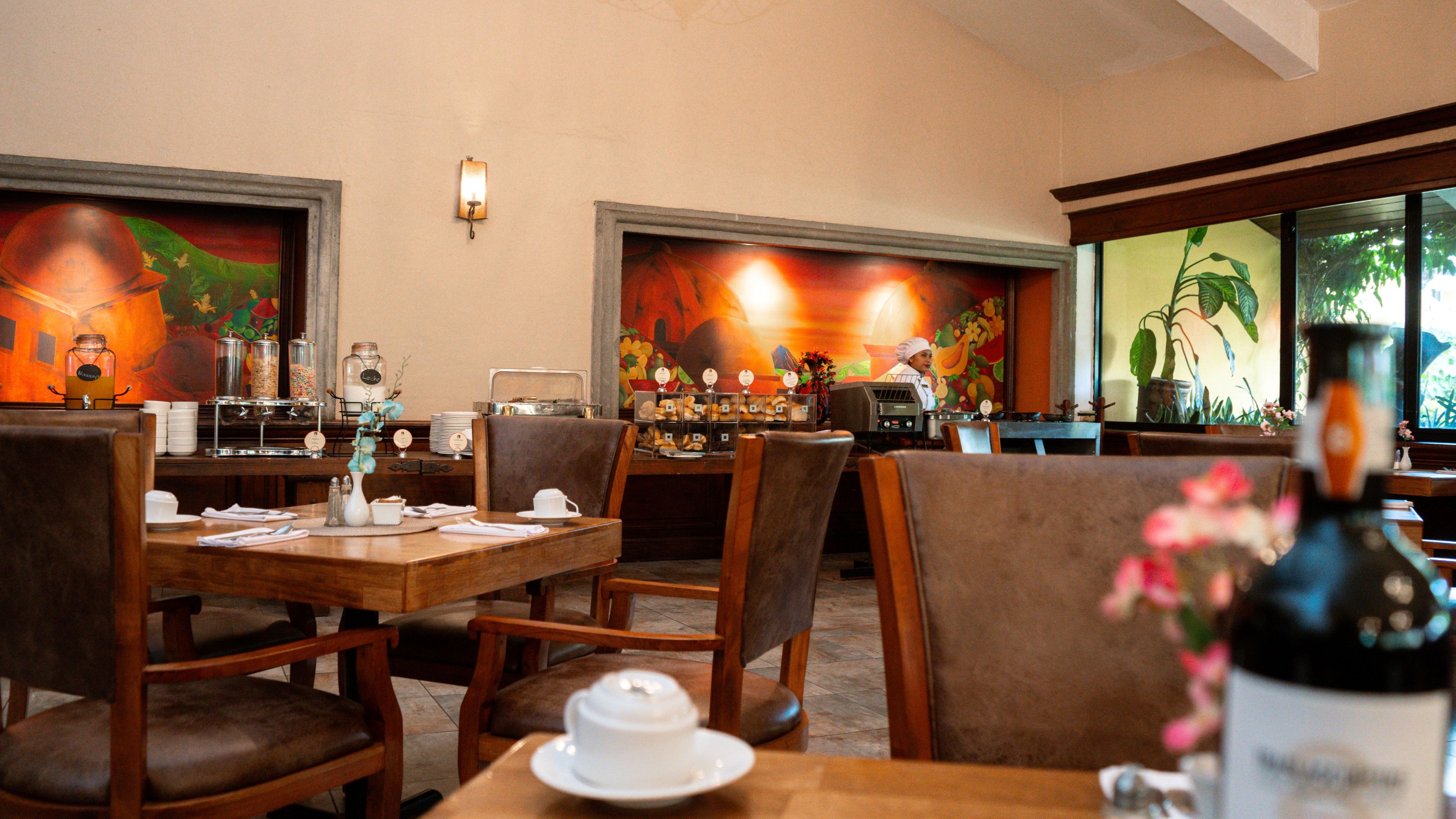A restaurant with wooden tables and chairs, a coffee station, and a mural on the wall.