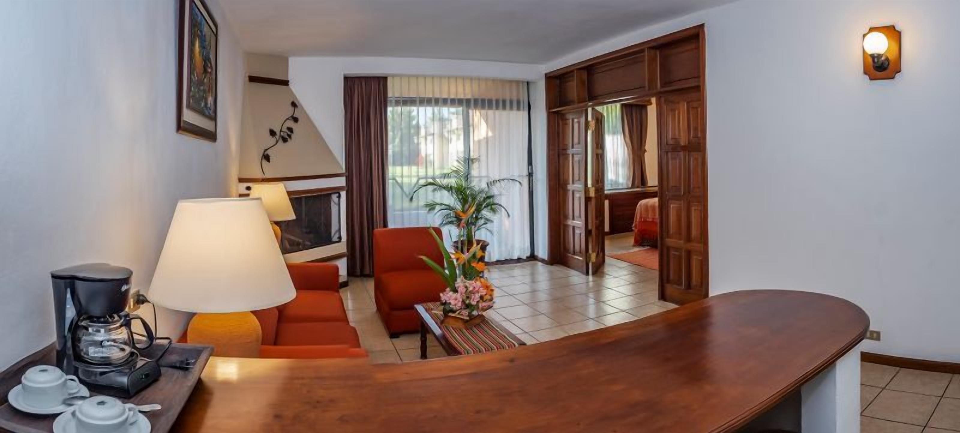 A hotel room with a wooden desk, a sofa, a coffee maker, and a lamp.