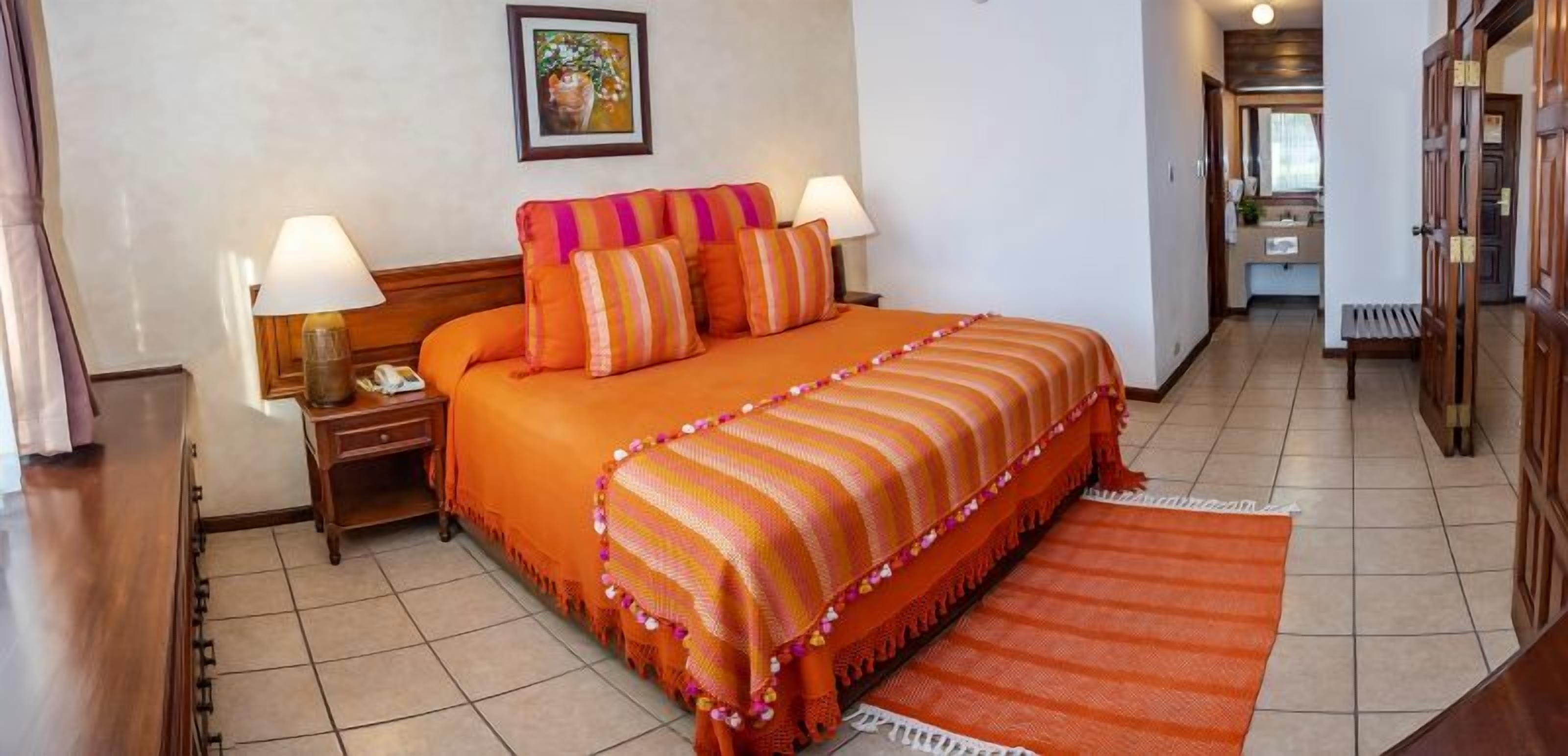 Une chambre d’hôtel avec un grand lit, deux tables de chevet, une commode, une armoire en bois et une salle de bain visible à travers une porte ouverte.