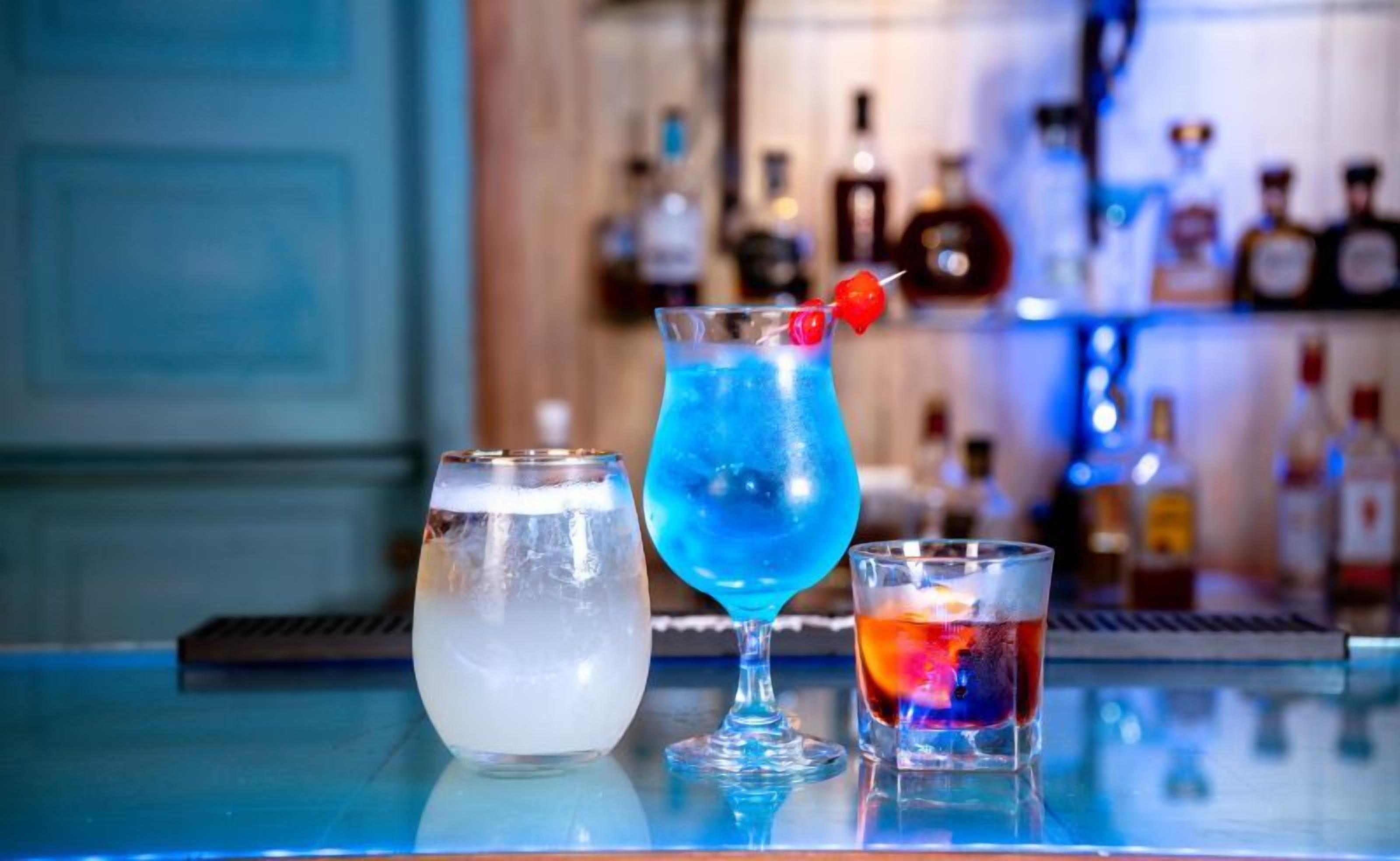 Trois verres contenant différentes boissons sont posés sur le comptoir d’un bar, avec en arrière-plan, flou, des étagères remplies de bouteilles.