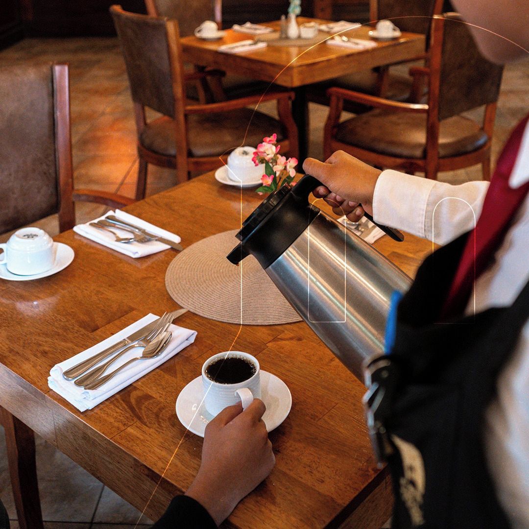 A person pouring coffee into a cup on a wooden table with a placemat, surrounded by chairs and a small table with a teapot and cups.