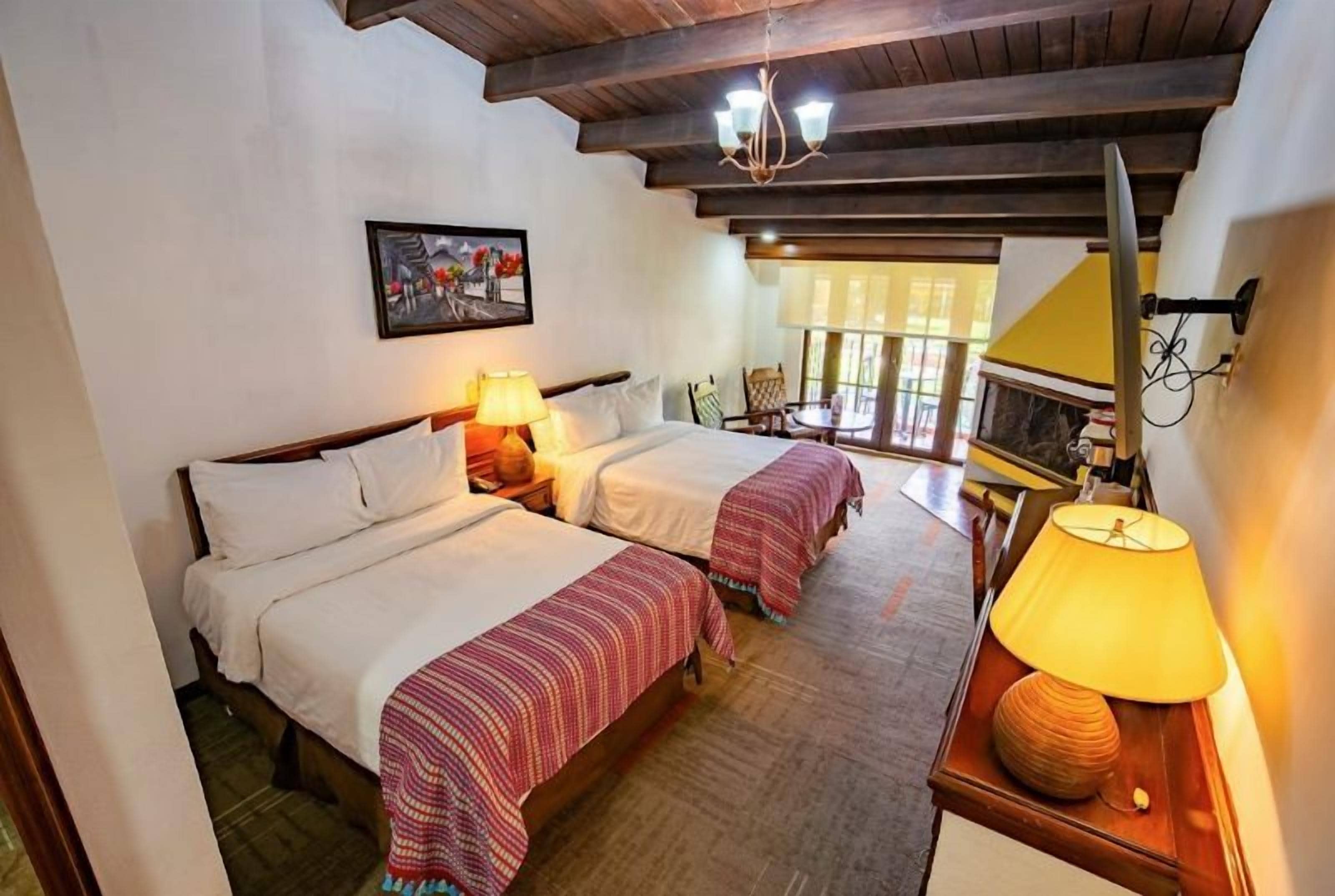 A room with two beds, a wooden ceiling, a table with a lamp, and a window with a view.