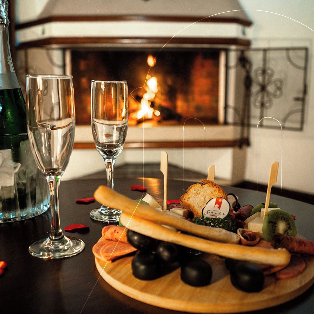 A wooden board with a variety of cheeses, olives, and a baguette, accompanied by two glasses of water, set on a table with a fireplace in the background.