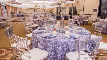 Un salón de banquetes con mesas redondas, sillas traslúcidas y tela blanca cubriendo la decoración.