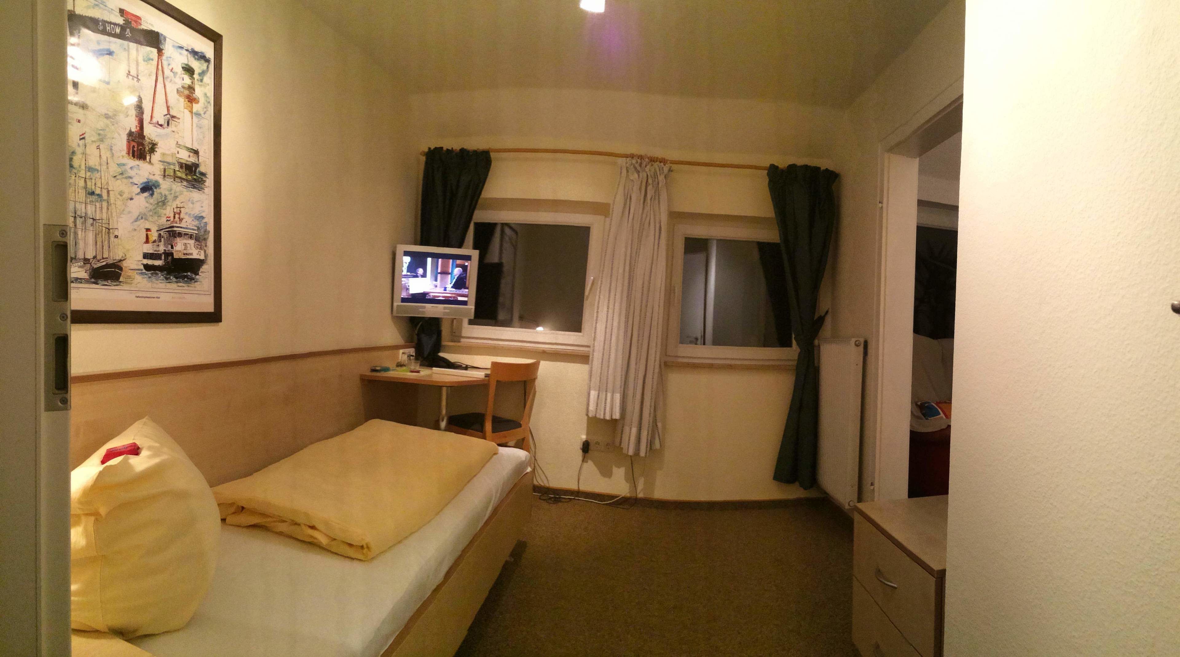 Une petite chambre d’hôtel avec un lit, un bureau avec un ordinateur, une fenêtre avec des rideaux et un mur orné d’un tableau.