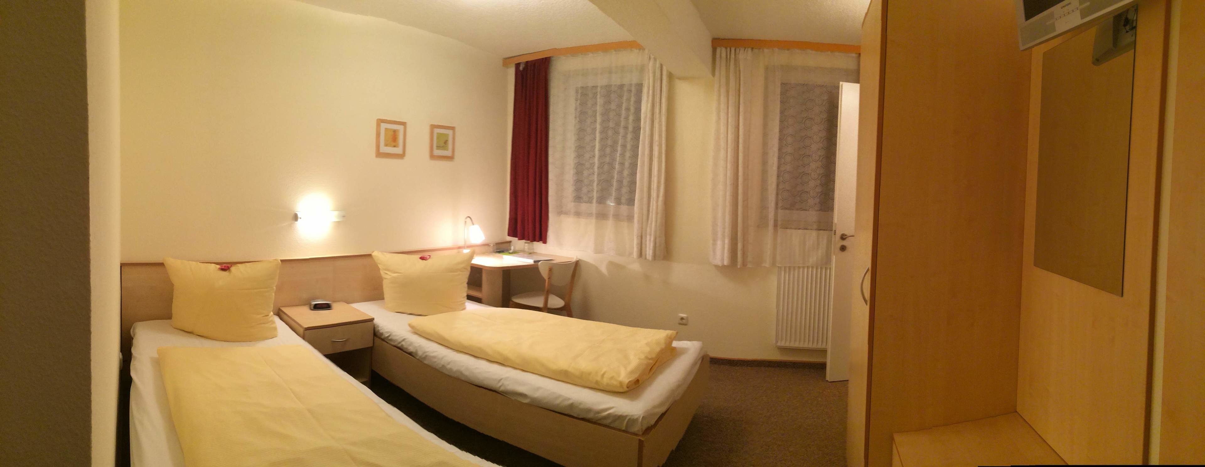 Una habitación de hotel con dos camas individuales, un escritorio pequeño con una silla, una ventana con cortinas y una puerta.