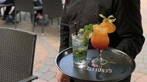 A person holding a tray with two drinks, a glass with a lime and mint garnish, and a glass with a straw and a slice of orange. - Hotel zur Heidebluete (Hambuehren)