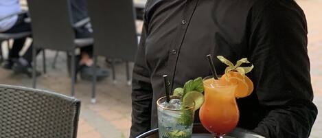 A person holding a tray with two drinks, a glass with a lime and mint garnish, and a glass with a straw and a slice of orange.