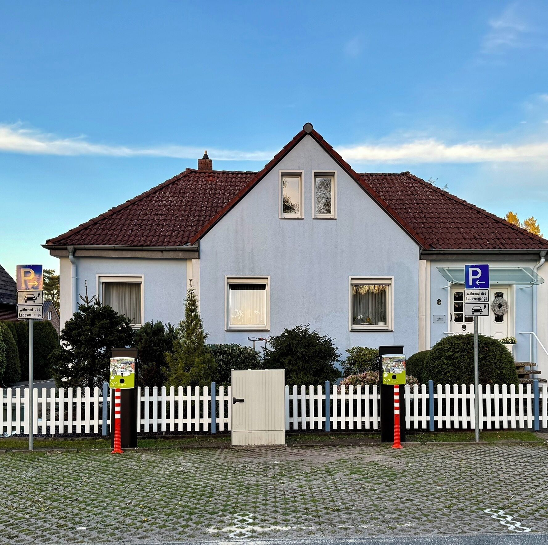 Ein zweigeschossiges Gebäude mit rotem Ziegeldach, weißem Zaun und einem Parkschild, das einen Parkplatz kennzeichnet.