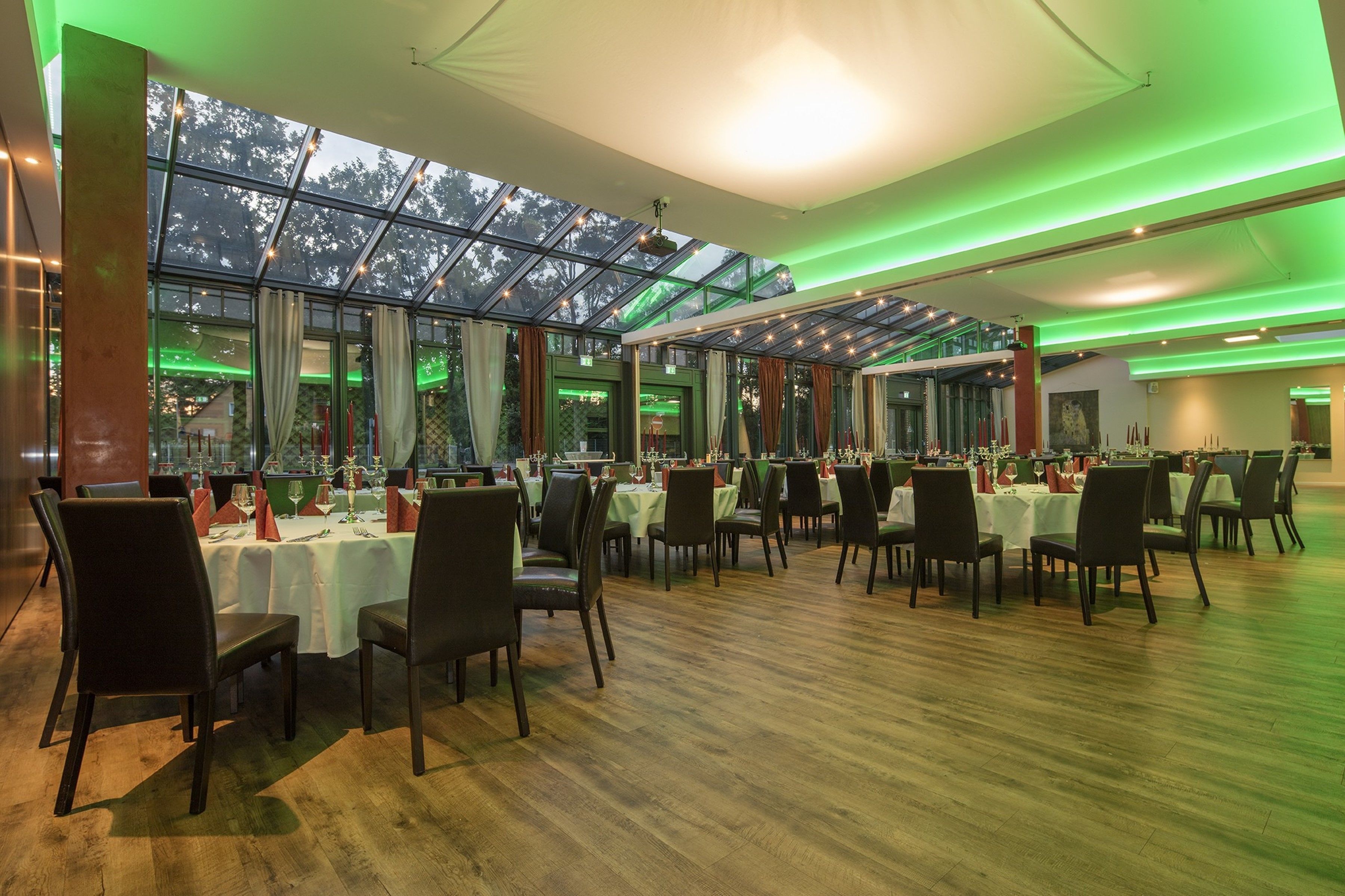 A spacious restaurant with multiple tables and chairs, large windows, and green ambient lighting.