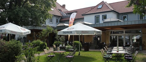 An outdoor seating area with tables and chairs under umbrellas, surrounded by greenery and buildings with a tiled roof.