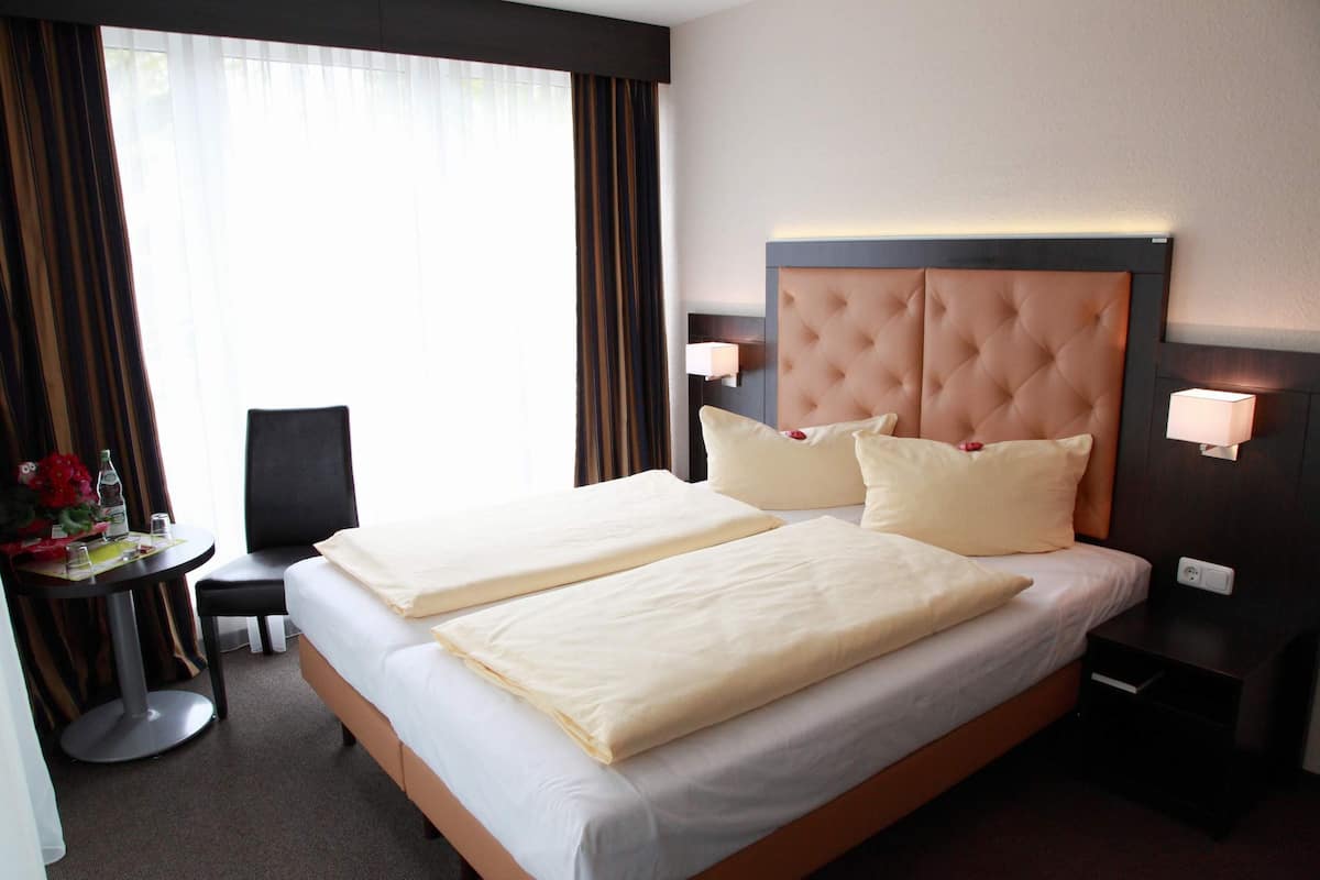A double bed with a tufted headboard, two bedside tables with lamps, a small table with a chair and flowers, and a window with curtains.