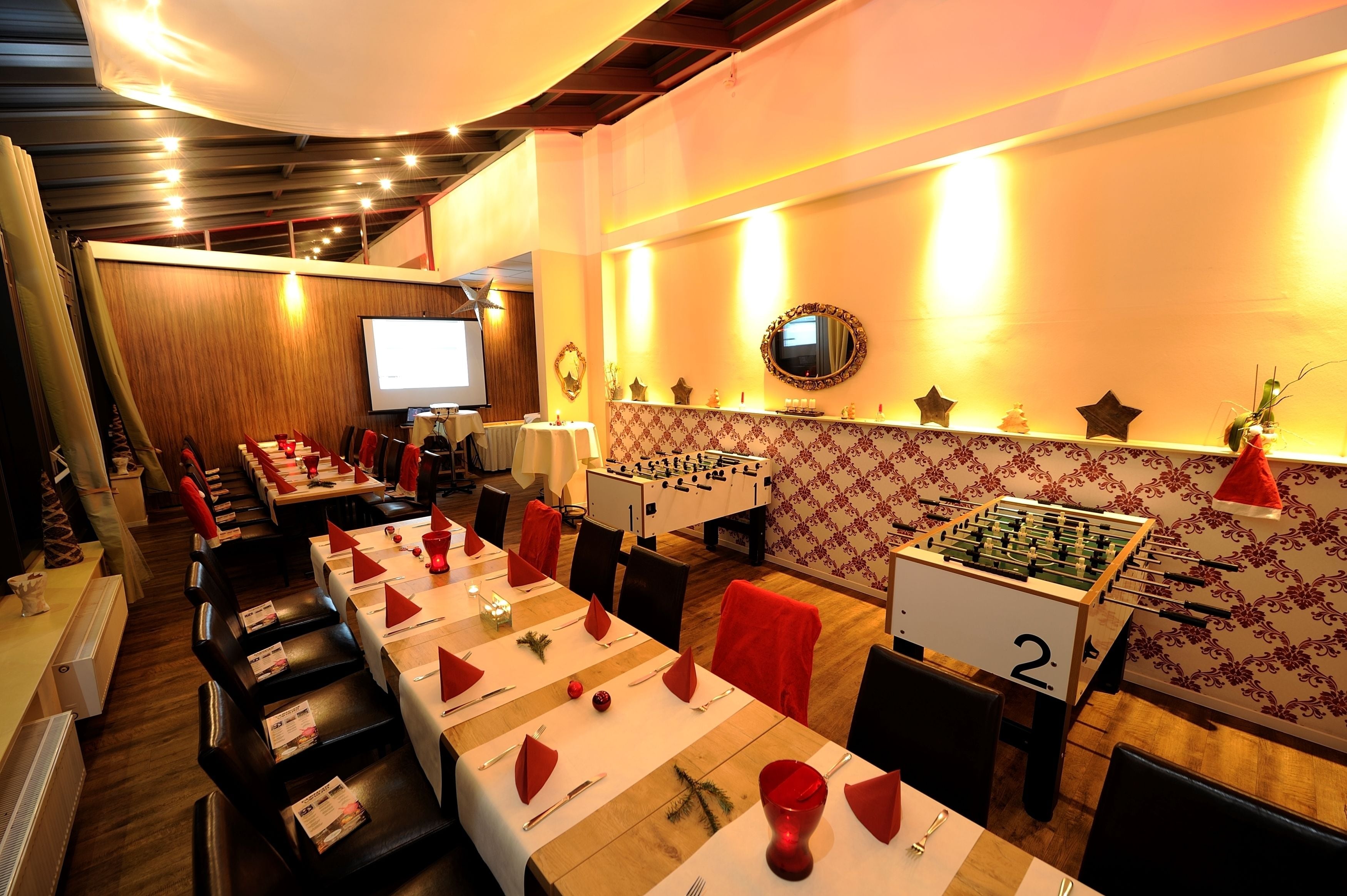 Un restaurant avec de longues tables dressées pour le repas, un baby-foot et un écran de projection à l’arrière-plan.