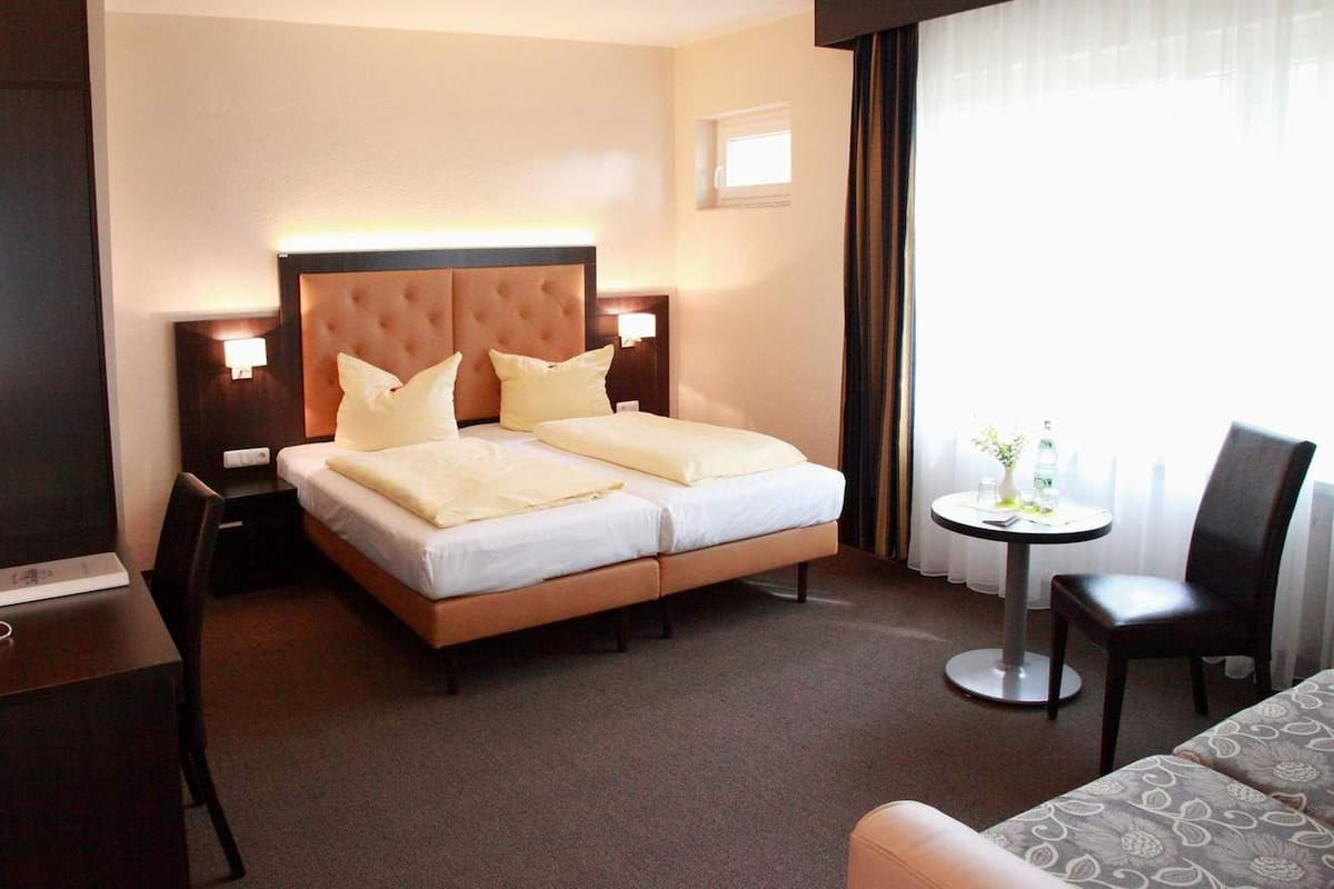 A hotel room with a double bed, bedside tables, a small round table with a chair, and a large window with curtains.