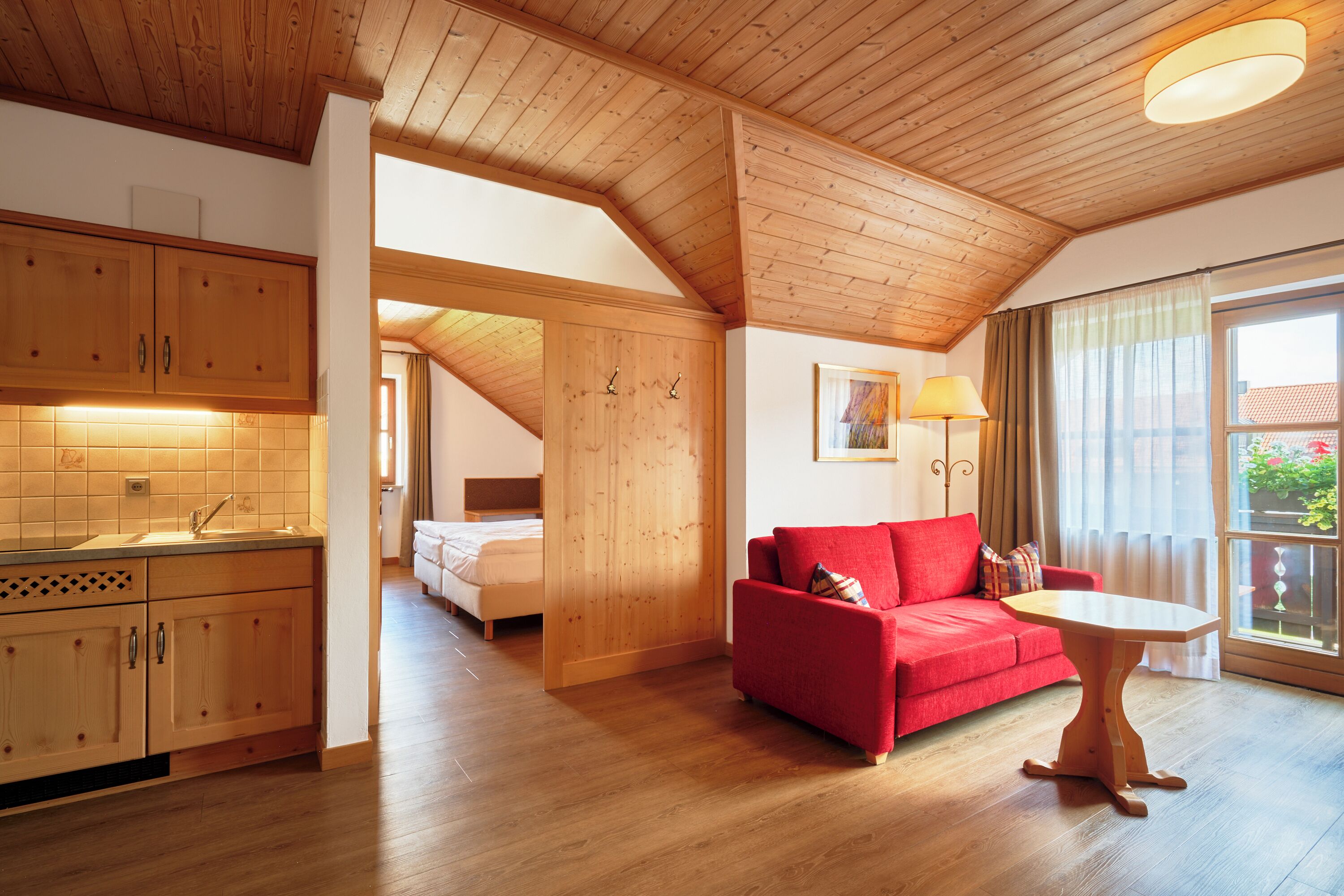 Ein geräumiger Raum mit Holzbalkendecken und -wänden, einem roten Sofa, einem kleinen Tisch und einem Fenster mit Vorhängen.