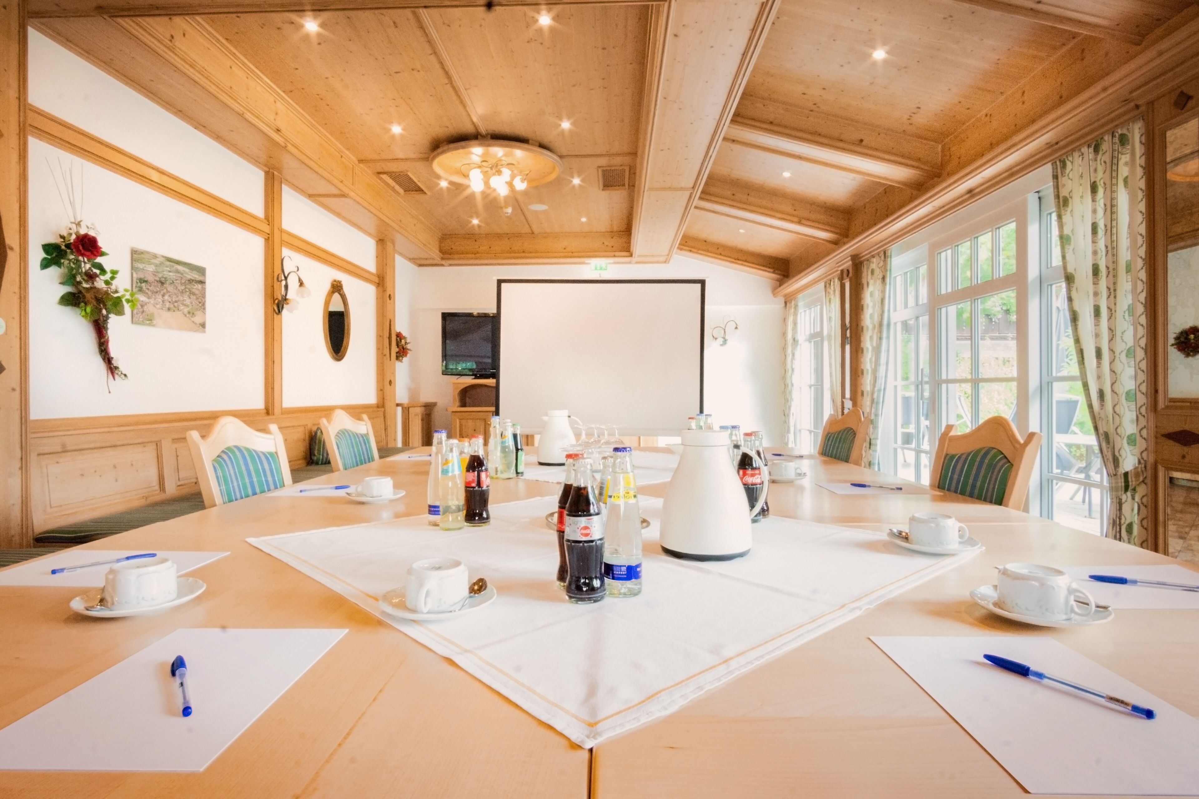 A conference room with a long table, chairs, a projector screen, and various items like cups, bottles, and a kettle.