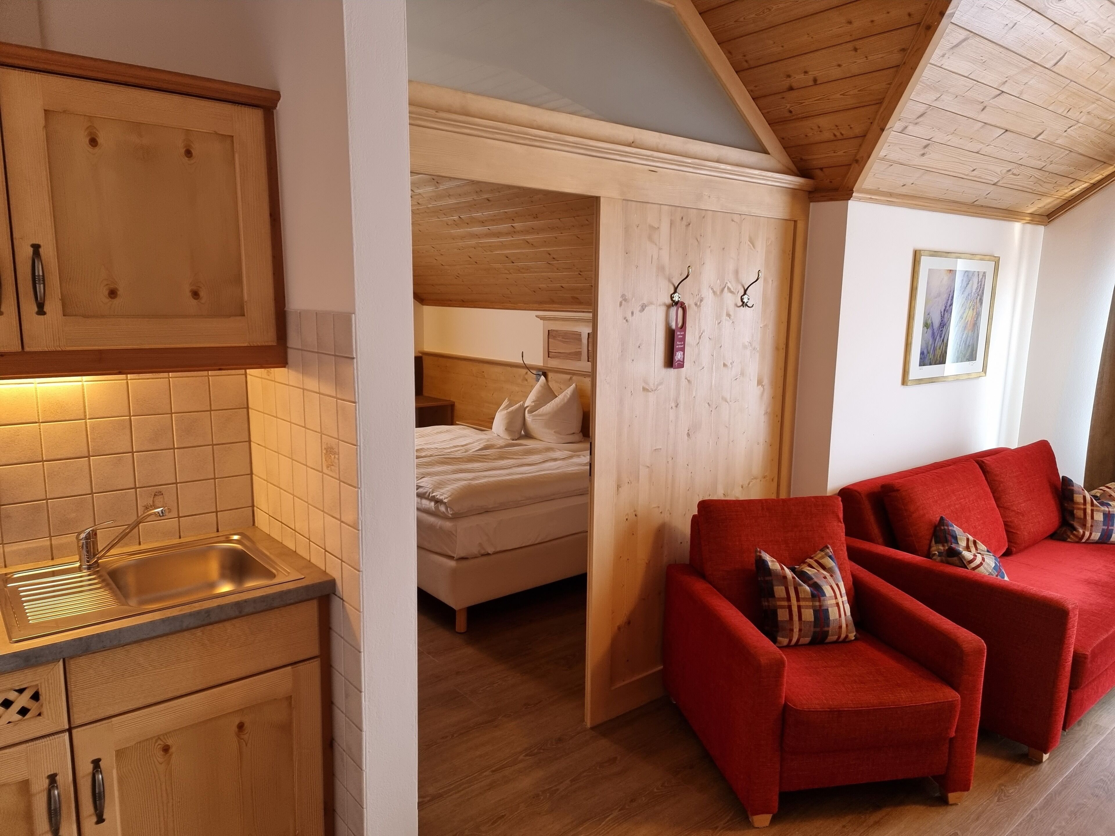 Een kleine kamer met een kitchenette, een bed en een rode slaapbank.