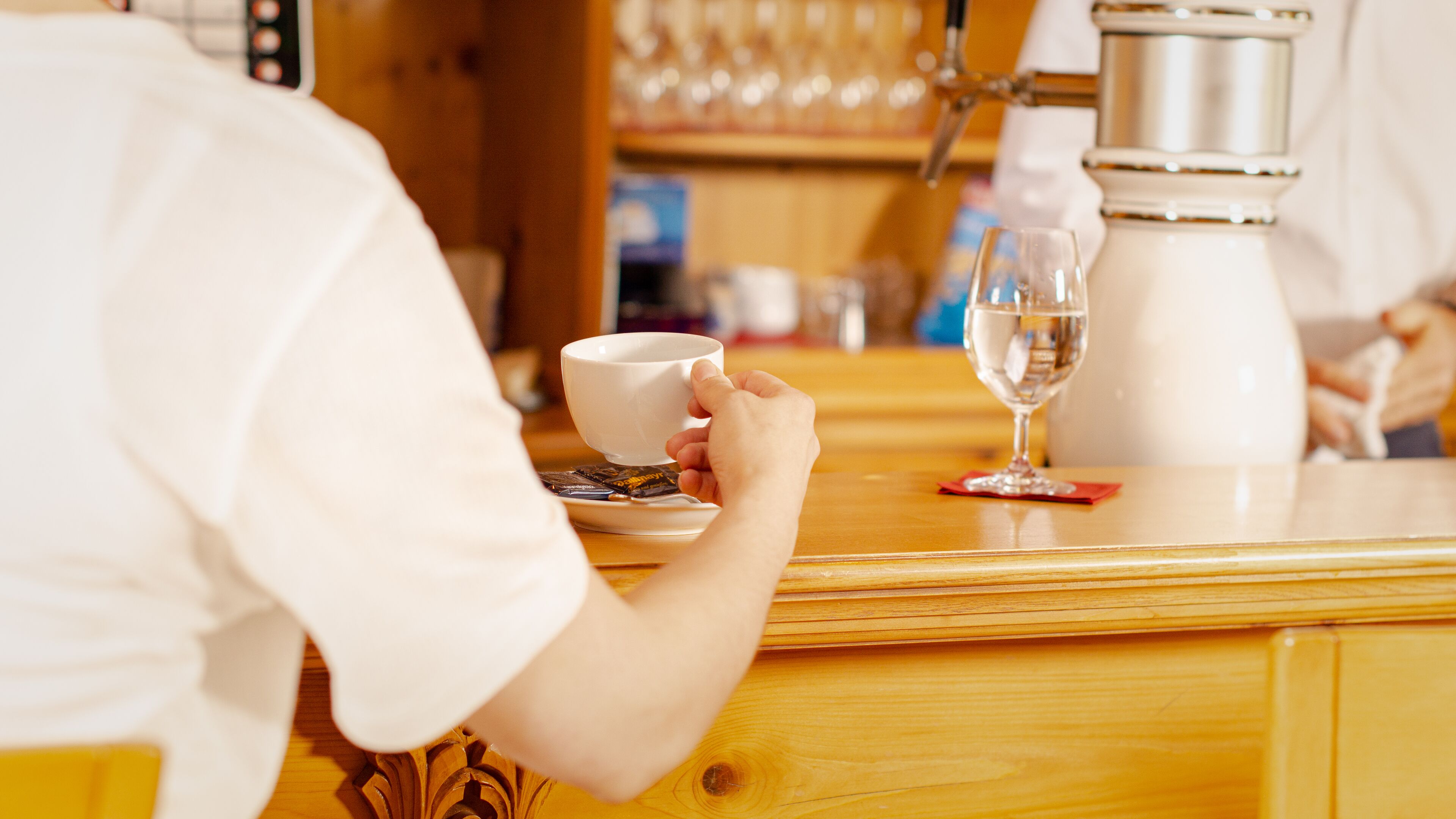 En person som håller i en mugg vid bardisken, där det också står en kaffemaskin och ett vinglas.