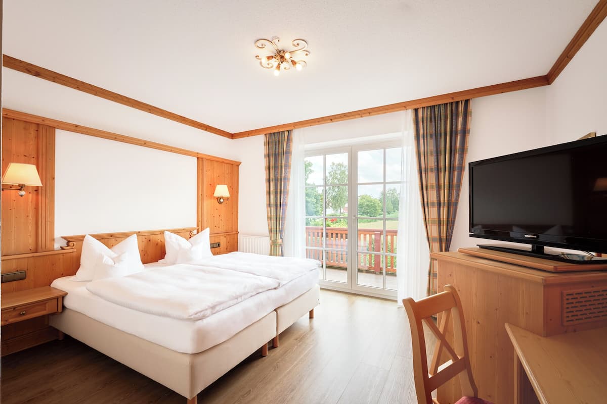 Quarto de hotel com uma cama grande, cabeceira de madeira, uma televisão sobre um móvel de madeira e vista para o exterior através de uma porta de correr de vidro.