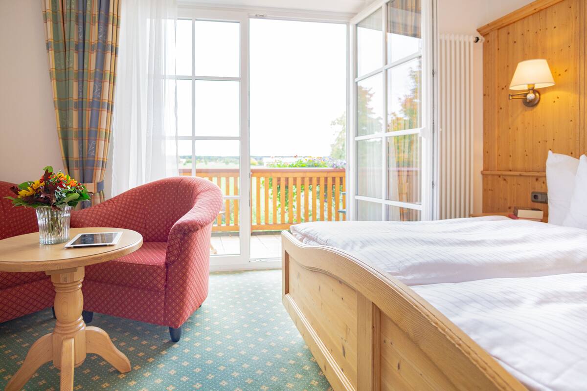 Quarto de hotel com cama, poltrona, mesinha com um vaso e um tablet, e vista para o exterior através de uma porta de correr.