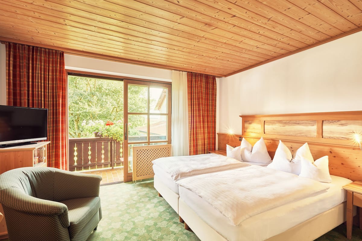 Quarto de hotel com teto de madeira, uma cama grande com roupa de cama branca, uma televisão sobre um suporte de madeira e vista para o verde lá fora.