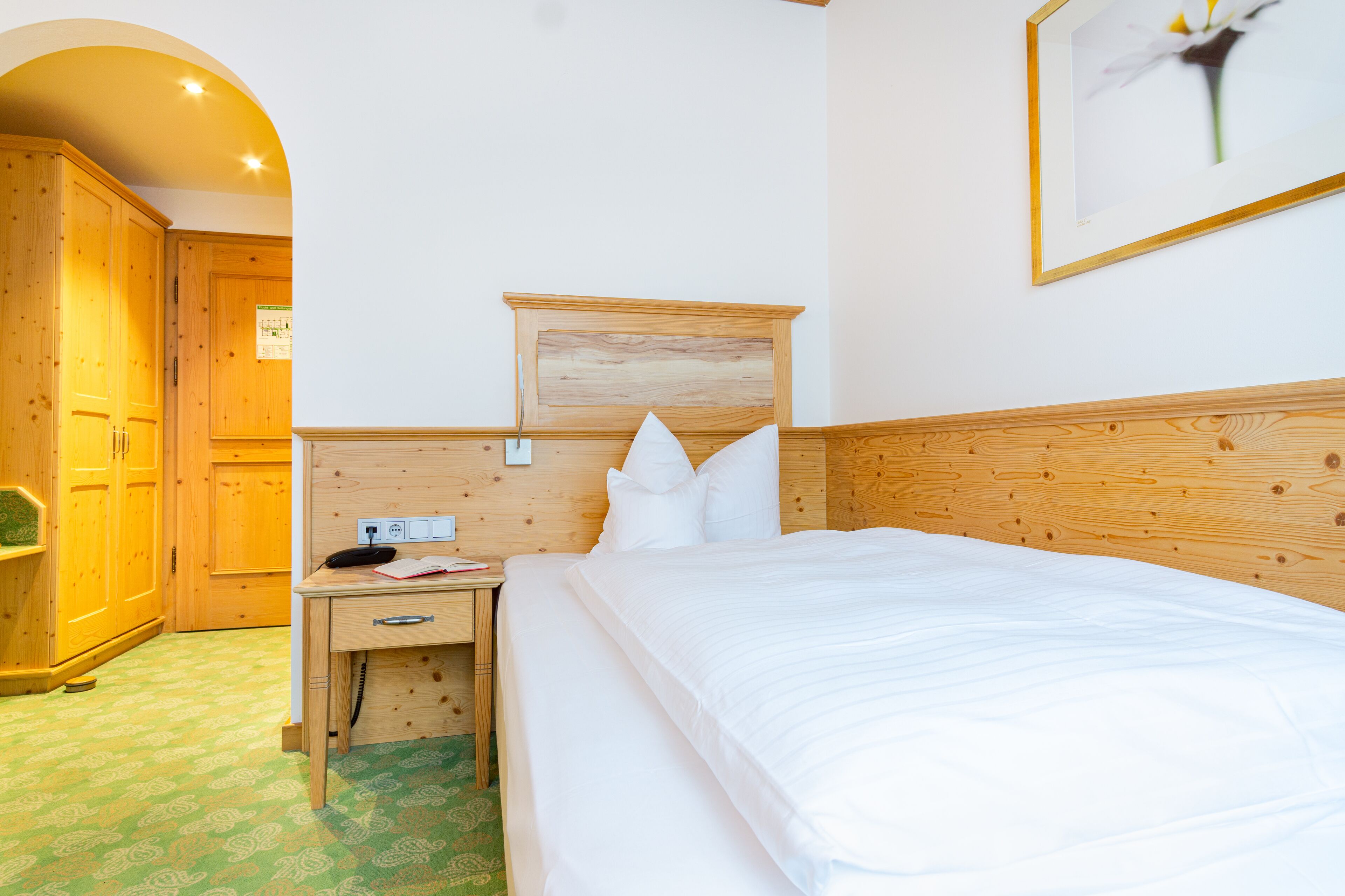 Habitación de hotel con cabecera de madera, cama con ropa blanca, mesita de noche con un teléfono y un libro, y un cuadro en la pared.