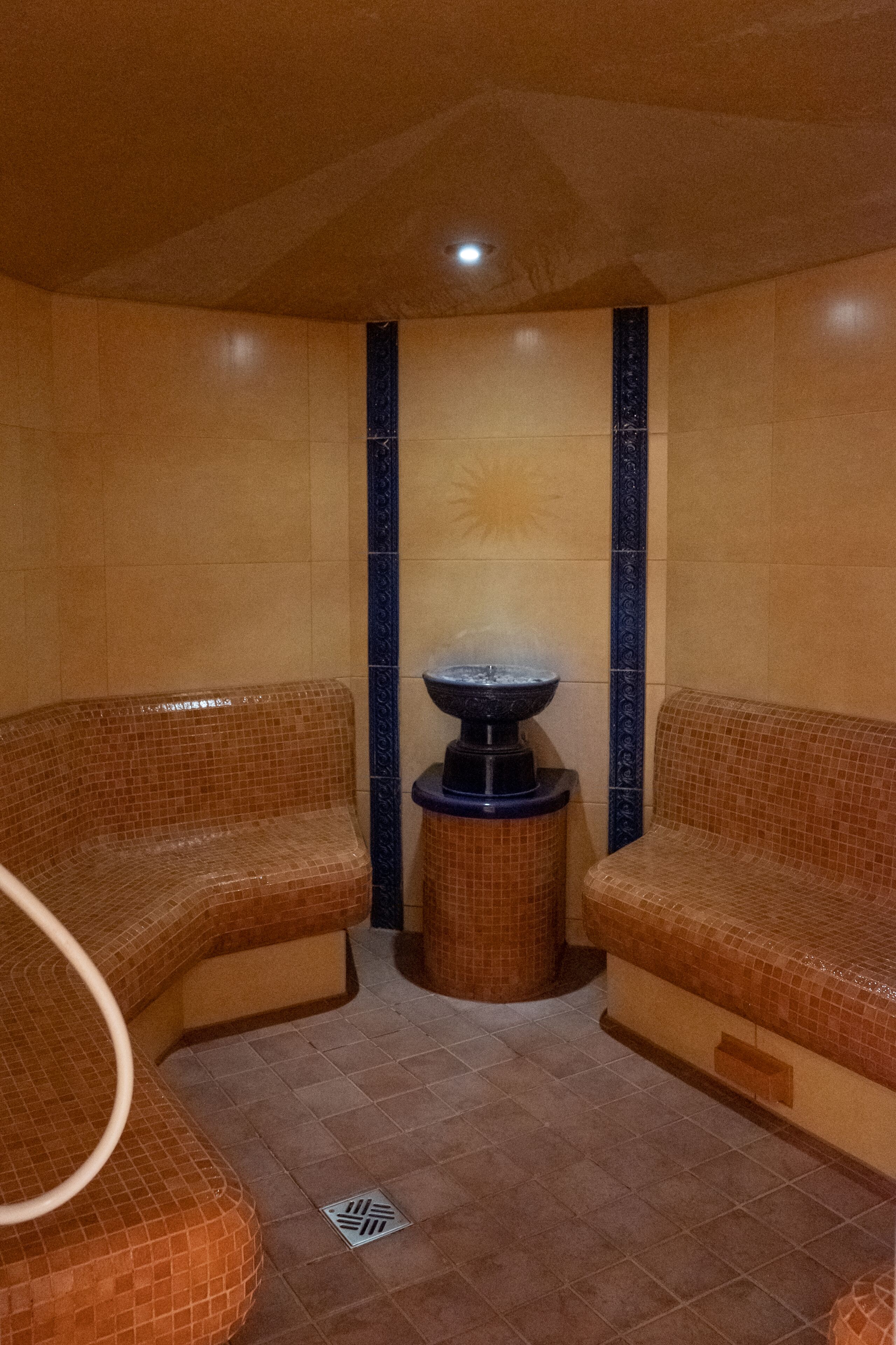 A spa room with two cushioned seating areas, a central water feature, and tiled walls and floor.