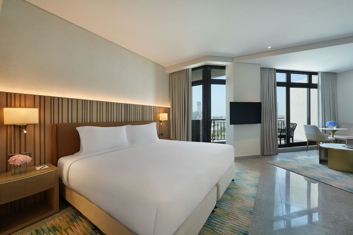 Ein Hotelzimmer mit einem großen Bett, einem Schreibtisch mit Lampe, einem Fernseher und Blick auf die Stadt durch große Fenster.