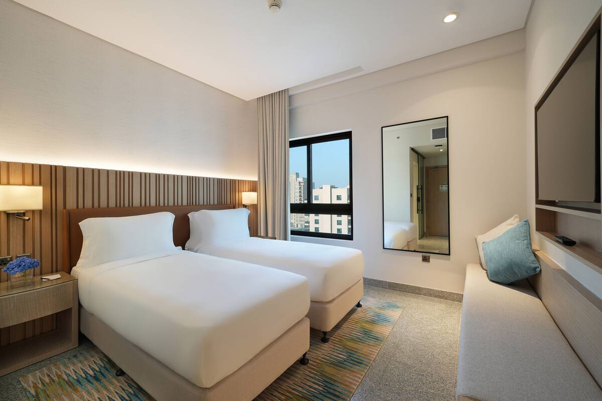 Ein Hotelzimmer mit zwei Betten, einem Nachttisch, einem Schreibtisch, einem Spiegel und einem Fenster mit Blick auf die Stadt.