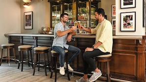 Dos hombres brindando con bebidas en un bar con taburetes de madera y lámparas colgantes.