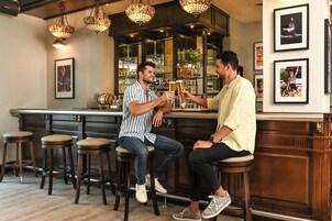 Two men toasting with drinks at a bar with wooden stools and hanging lamps.
