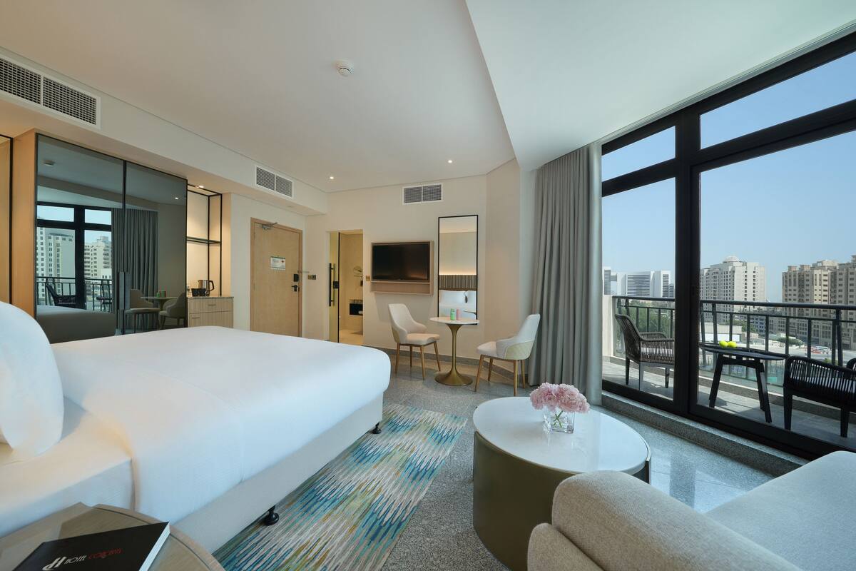 Ein Hotelzimmer mit einem großen Bett, einem Schreibtisch, einem Fernseher, einem Balkon mit Sitzgelegenheit und Blick auf die Stadt.